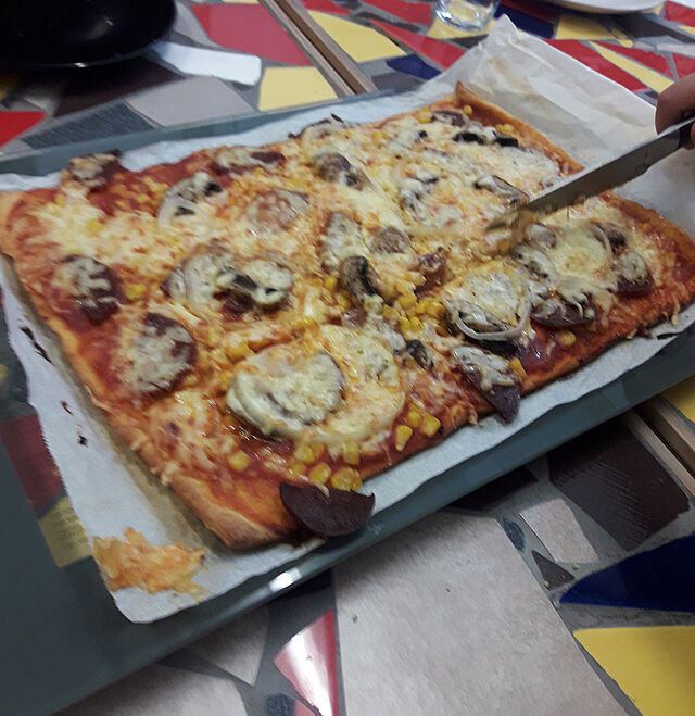 Eine fertige Pizza steht mit dem Blech auf dem Tisch.
