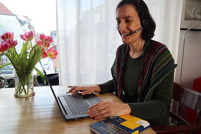 Eine Frau sitzt mit Headset und Laptop am Esstisch. Auf dem Tisch ein Strauß roter Tulpen.