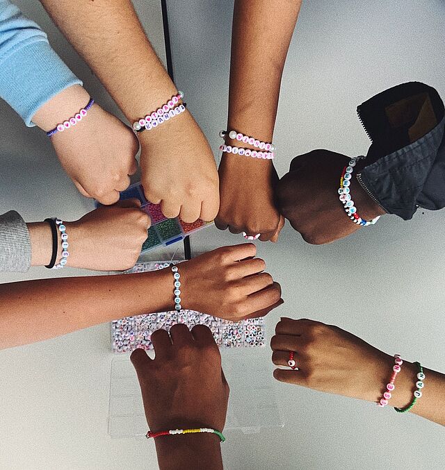 Acht Arme unterschiedlicher Hautfarbe und mit selbstgebastelten Armbändern geschmückt bilden einen Kreis. 