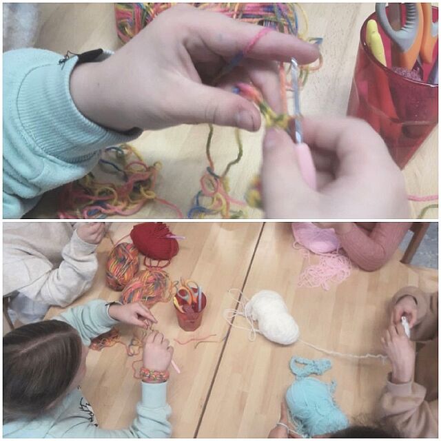 Es ist ein Tisch zu sehen, um den mehrere Kinder sitzen. Sie halten Häkelnadeln und Garn in der Hand. Auf dem Tisch liegen Wollknäule.