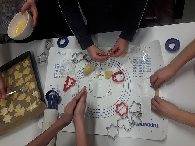 Mehrere Kinderhände stechen Kekse aus. Eine andere Hand bestreicht Kekse auf einem Blech mit Ei.