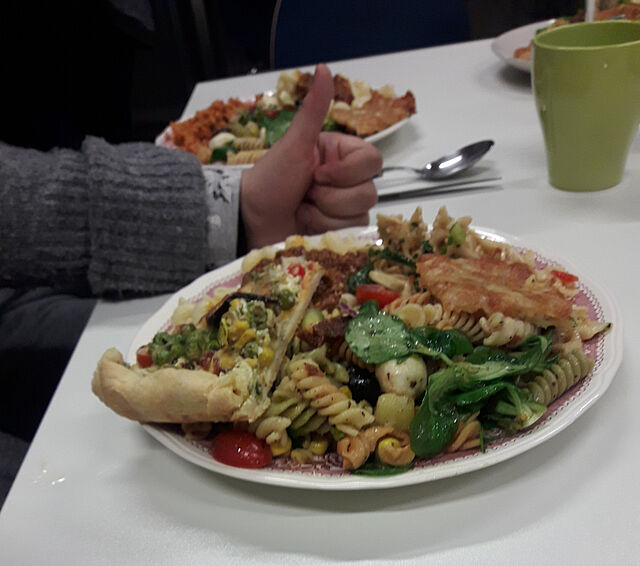 Neben einem vollen Teller liegt eine Hand und zeigt mit dem Daumen nach oben, dass es lecker ist.