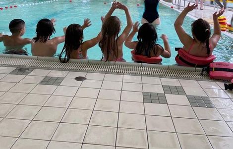 Sechs Kinder am Schwimmbeckenrand im Wasser mit dem Rücken zur Kamera und halten die Hände in die Höhe