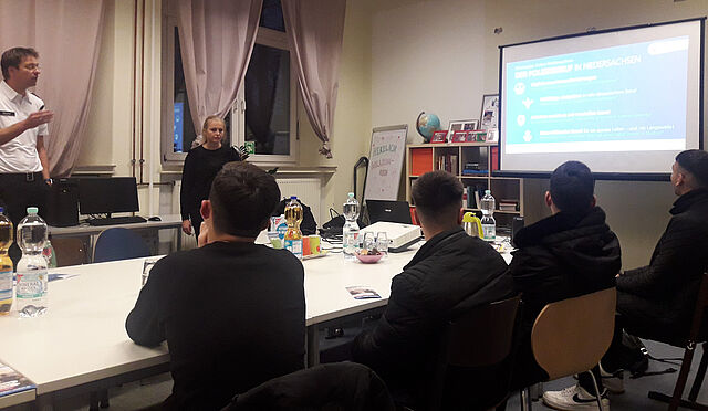 Vier Jungen hören einem Mann und einer Frau zu, die zu einer PowerPointPräsentation sprechen.