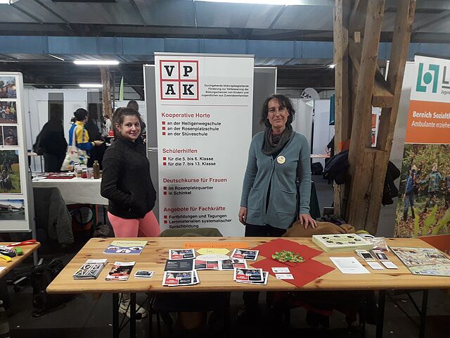 Hinter einem Tisch mit Informationsmaterial des VPAK e.V. stehen zwei Frauen.