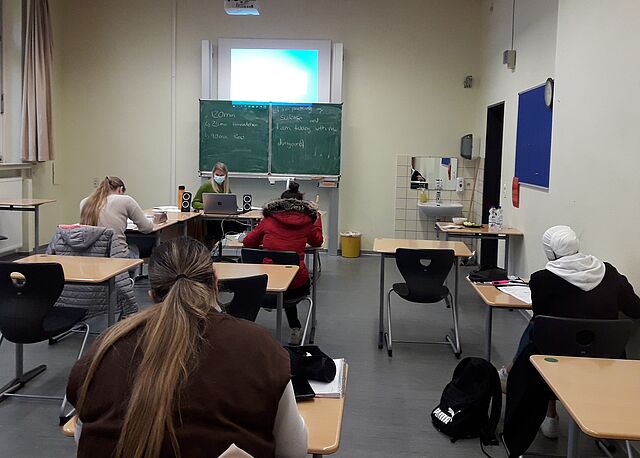 Jugendliche sitzen in einem Klassenzimmer und folgen dem Unterricht einer Lehrerin. 