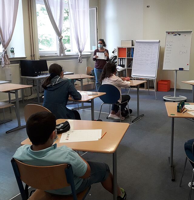 Jugendliche sitzen an Einzeltischen im Klassenraum und hören einer Lehrkraft zu.