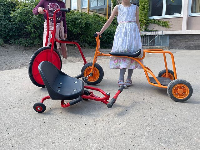 Zwei Kinder stehen draußen hinter einem orangenen Dreirad, einem roten Hochrad und einem roten Racer.