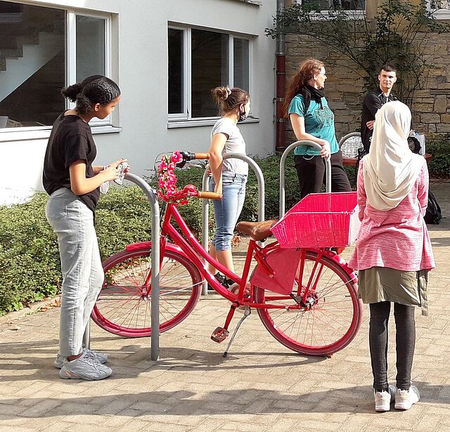 Vor einem Schulgebäude stehen drei Jugendliche und hören aufmerksam dem FSJ-ler und der Projektleitung zu.